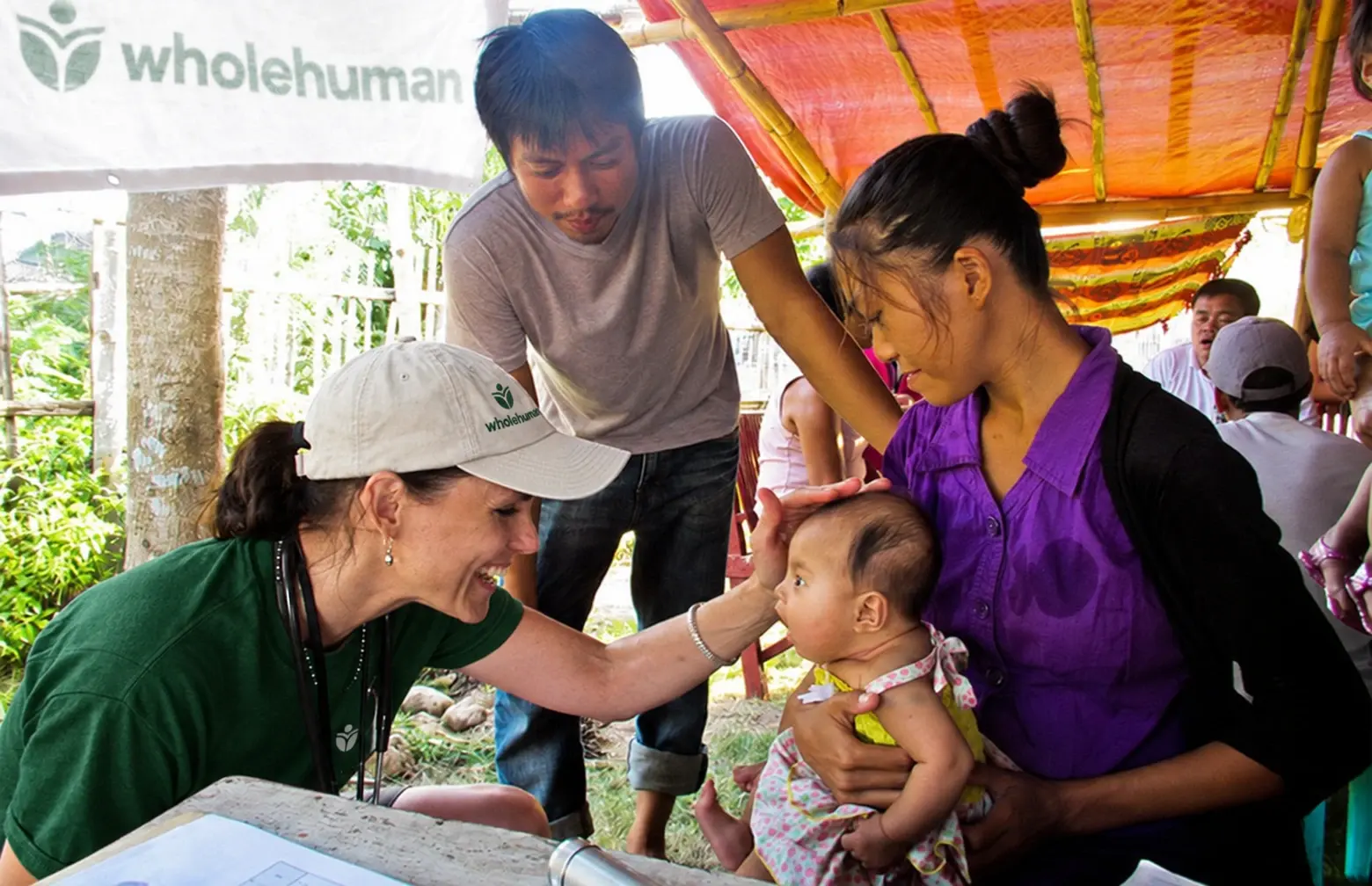 A Wholehuman team member wearing a stethoscope smiles at a baby held by her mother